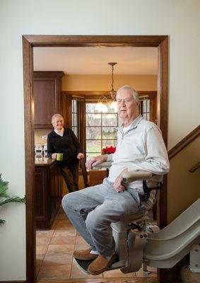 man riding curved stair lift in his home while walk holds coffee cup in background