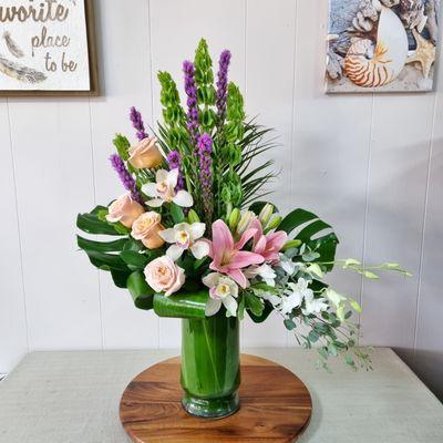 Designer Choice Arrangement with Bells of Ireland, Roses, Cymbidium Orchids, Dendrobium Orchids, Liatris, Pink lilies and tropical greenery.