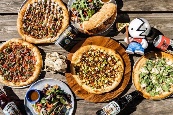 Outdoor picnic table on a sunny day with pizzas, salads, and drinks.