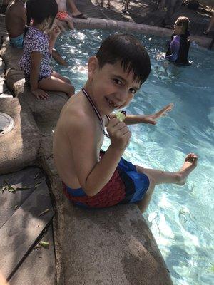 Happy kid sitting on the side of the pool, showing off his medal on the last day of swim lessons.