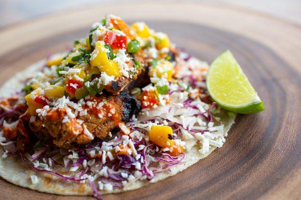 Tandoori-Style Chicken Taco. Grilled chicken marinated in yogurt, lemon, and masala spices, slaw, red chile crema, mango salsa, cotija.