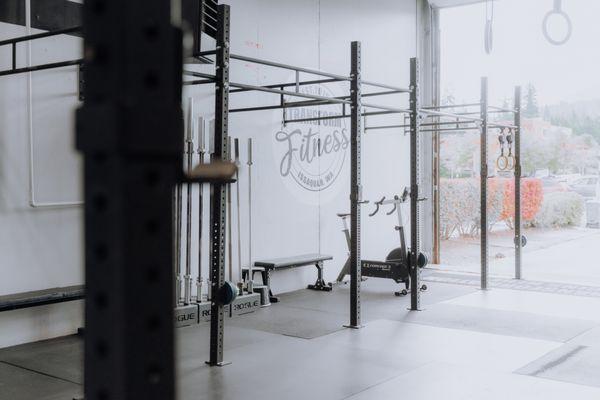 "A gym floor with Rogue racks along one side, holding barbells and weights.