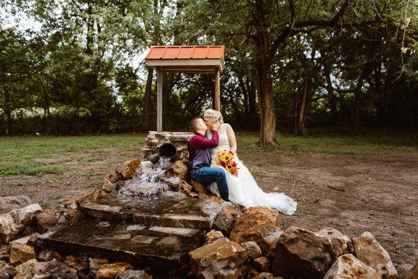 A wedding couple poses at the spring well with a stream of water falling down into the rock-lined creek below at the Spring Creek venue.