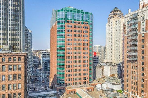 A vibrant cityscape featuring a tall, distinctive red-brick building with green-tinted windows and a modern geometric design....