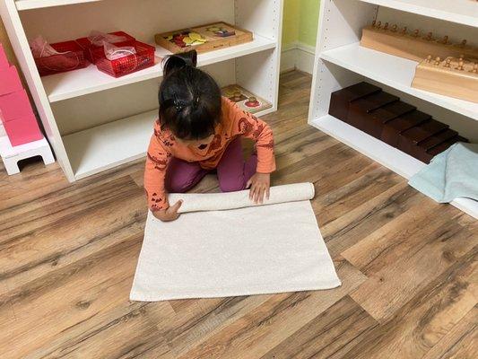 A two year old student rolling a rug using her fine motor skills after finishing her job.