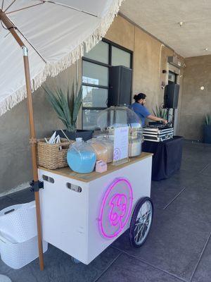 The cart is outside a place of business with a DJ in the background. The cart has the pink Neon sign and has blue and orange sugar in jars.