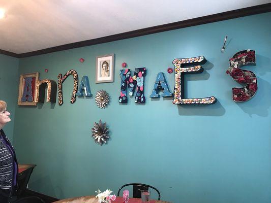 Spells "Anna Mae's." The letters are decorated with things like flowers, buttons, corks, bottle caps, and a mustache and glasses!