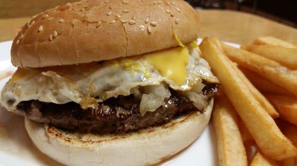 A Cheeseburger with fried onions and a fried egg on a toasted sesame seed bun with a side of fries.