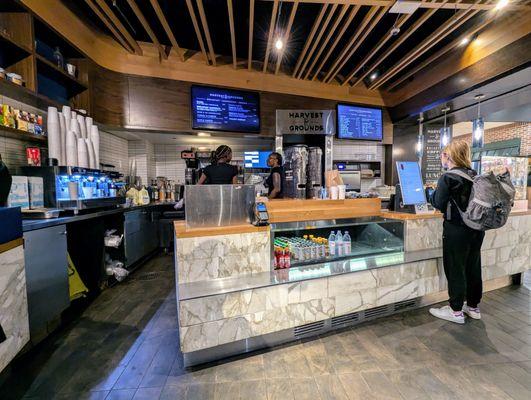 Inside Harvest & Grounds next to Gate D9. Open prep kitchen, self-service ordering kiosk touchscreen tablet on right. Waiting for my order.