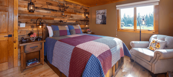 Nightstands flank a bed with a patchwork quilt and large armchair. A pallet wall and railroad lanterns are behind the bed.