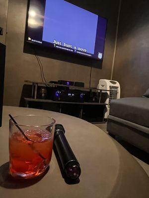 ID: In the foreground, a cocktail and microphone rest on a coffee table. On the TV screen are lyrics to "Bust A Move" by Young MC.