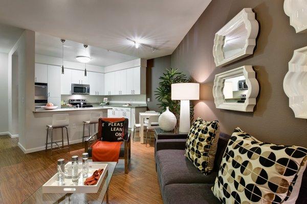 Staged living room with hardwood style vinyl flooring. Kitchen in the background with stainless steel appliances and breakfast bar.