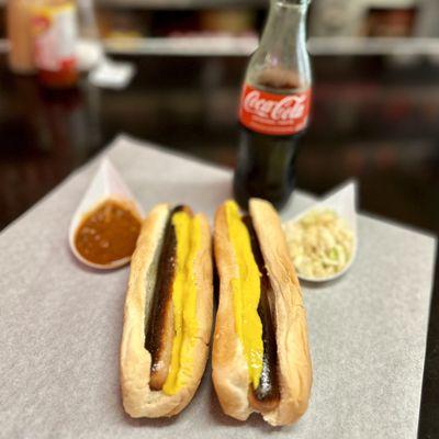 Two hot dogs with mustard. Glass Bottled Sodas. Chili and slaw on the side.