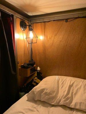 The little built in lamp, charging station and mini-nightstand surface inside my bunk. Note the blackout curtain and super fluffy pillow