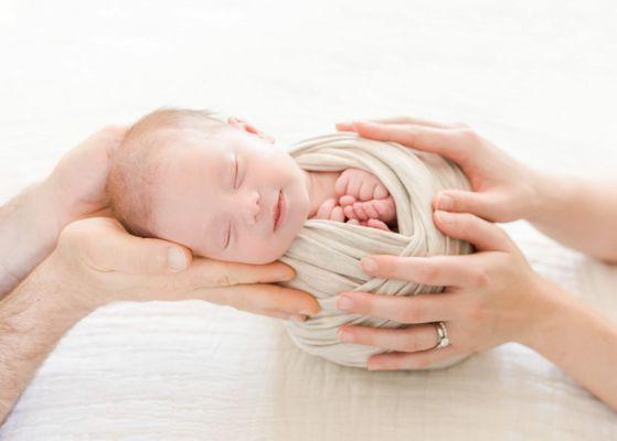 Parents' hands holding newborn baby in a cream wrap.