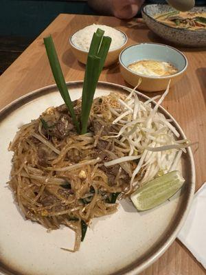 Beef pad thai with white rice, fried egg and curry in the background.