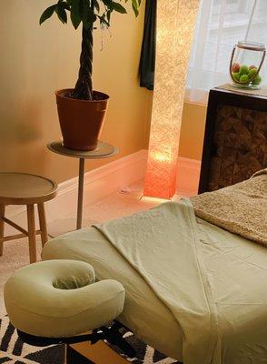 A massage table is made up with comfy sage green sheets. The massage studio is in a palette of greens and browns, very relaxing.