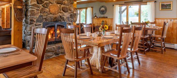 Wooden table with tree stump base in front of large stone fireplace with another table near a large window.