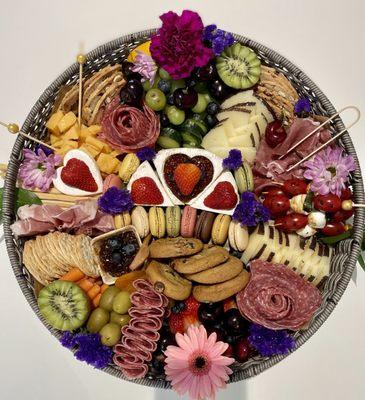 This round rattan basket tray 16X16 filled with array of cheeses, cured meats, seasonal fruits and crudités, and crackers.