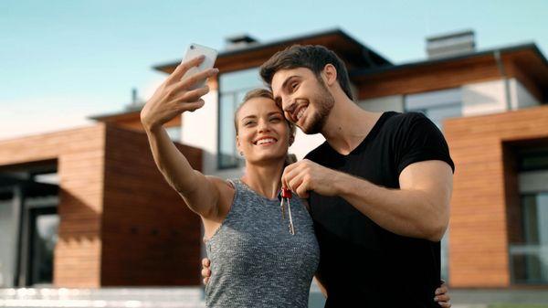 Young couple taking a photo with their phones in front of their new home.