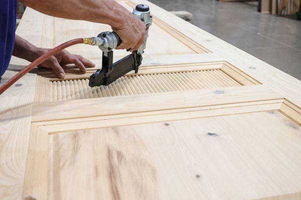 A close-up of a person's hands using a pneumatic nail gun to assemble a wooden door. The door features a decorative louvered ...