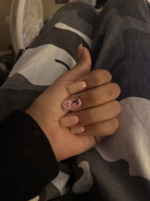 Square Nude nails with a light pink on the middle finger with a black A and a white heart