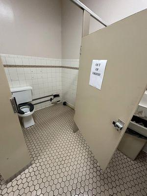 An accessible toilet stall, with a sign saying out of order. There is a hole in the wall where some plumbing is missing.