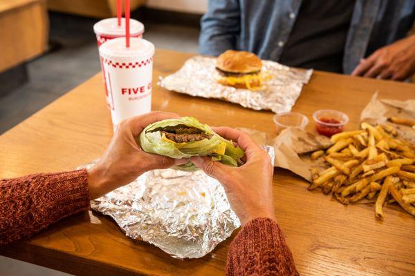 A customer's hands hold a Five Guys lettuce wrap at a dining room table inside a Five Guys restaurant.