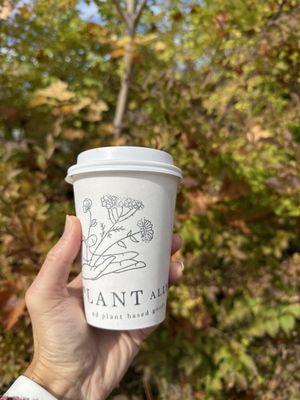 A hand holding a recyclable paper coffee cup with The Plant Ally written on the side below the drawing of a hand holding flowers