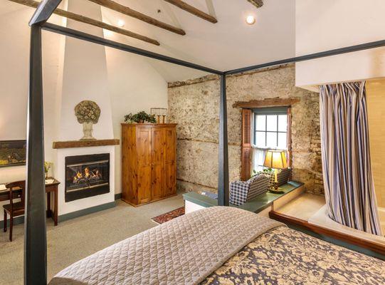 Corner of historic room with fireplace, wardrobe, and sitting area by window seen over corner of a comfortable 4-poster bed