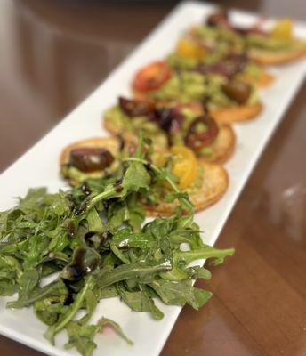 Avocado Toast, a harmonious blend of smashed avocado, heirloom tomatoes, arugula, pickled onion, and EVOO on garlic toast.