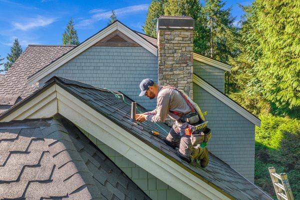 A skilled roofing contractor wearing safety gear works diligently on an asphalt shingle roof, performing repairs around a roof vent.