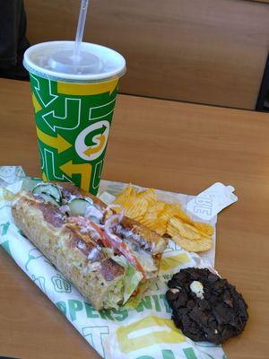 A buffalo chicken sub with lettuce, tomato, red onion, cucumber, and ranch dressing. A side of chips and a double chocolate chip cookie