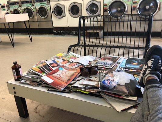 Coffee table flooded with magazines and topped with trash. Individual chairs have been replaced with industrial bench seating.