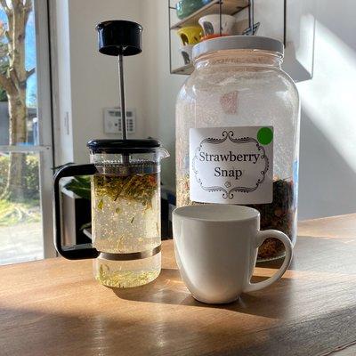 A French press infuses tea in a sunbeam. There is a white mug beside it and a jar of tea leaves labeled "strawberry snap."