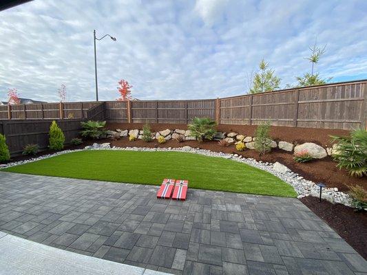 Clean and simple backyard.  Artificial grass with patio. Palm trees, mountain hemlock, various shrubs.