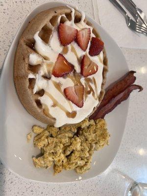 Gingerbread waffle with homemade whipped cream, maple syrup, fresh cut strawberries, cheesy eggs, and bacon.