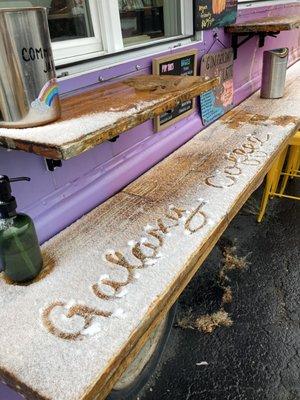 The words "Galaxy Coffee" written in the snow that is lightly dusted on the wooden counter attached to The Galaxy Coffee truck