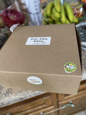 Cardboard cake delivery box with "Crum Cakes Bakery, made with care," "Happy Birthday!", and "You've got great taste" stickers.