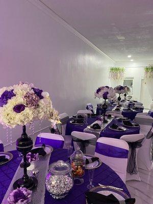 Purple tablecloth with purple tie backs and silver runner. Custom vase with floral centerpiece.