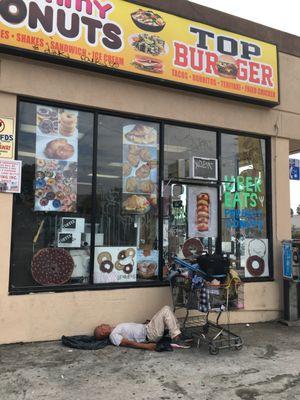 A man with a shopping cart snoozes in front of Yummy Donuts/Top Burger.