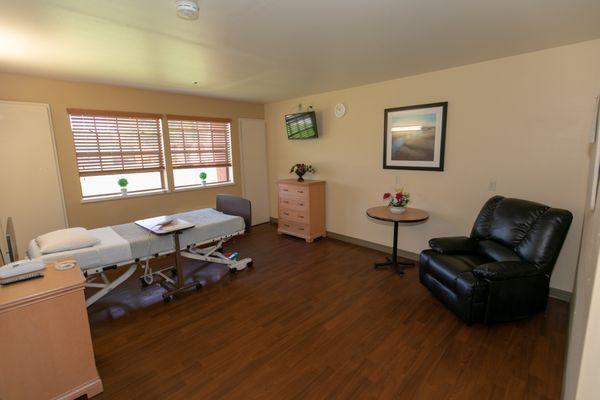 Room includes: bed, bedside table, call button, television, remote, table, seating, dresser, night stand, closet and bathroom.