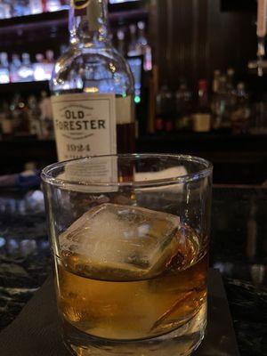 Bottle of Old Forester bourbon and a glass of bourbon. In the background are bottles of bourbon.