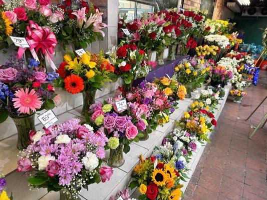 Vibrant flower arrangements on display in an open-air market, featuring roses, lilies, and sunflowers with festive ribbons and a balloon.