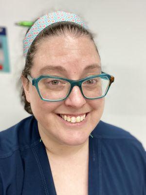 A white passing woman wearing teal semi-cat eye glasses and a navy blue top with a blue and pink patterned headband.