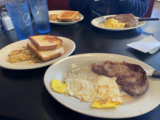 Rib-eye combo w/ eggs fried hard, sourdough toast, & hash browns. In back, t-bone combo w/scrambled eggs, sourdough toast, & hash browns.