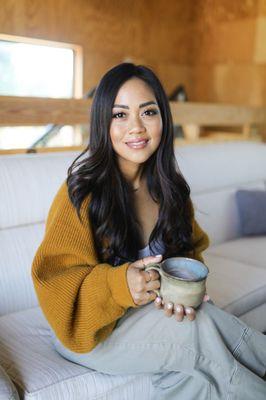 Monica Ramil is smiling while sitting on a couch holding a coffee mug