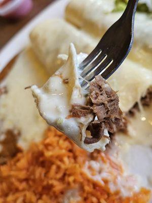 A piece of a Shredded Beef Chimichanga being taken by a fork, with the two Chimichangas, red rice, and refried beans are in the background.