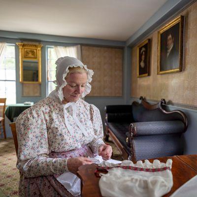 A woman in 1830s dress sews in the Fitch House