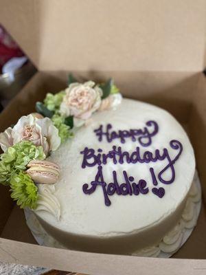 White birthday cake with purple "Happy Birthday, Addie!" icing message, live pink and green flower, macaron, and icing decorations.